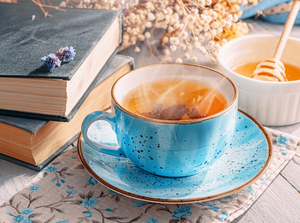 Tea in a light blue tea cup surrounded by flowers, books, and a bowl of honey.