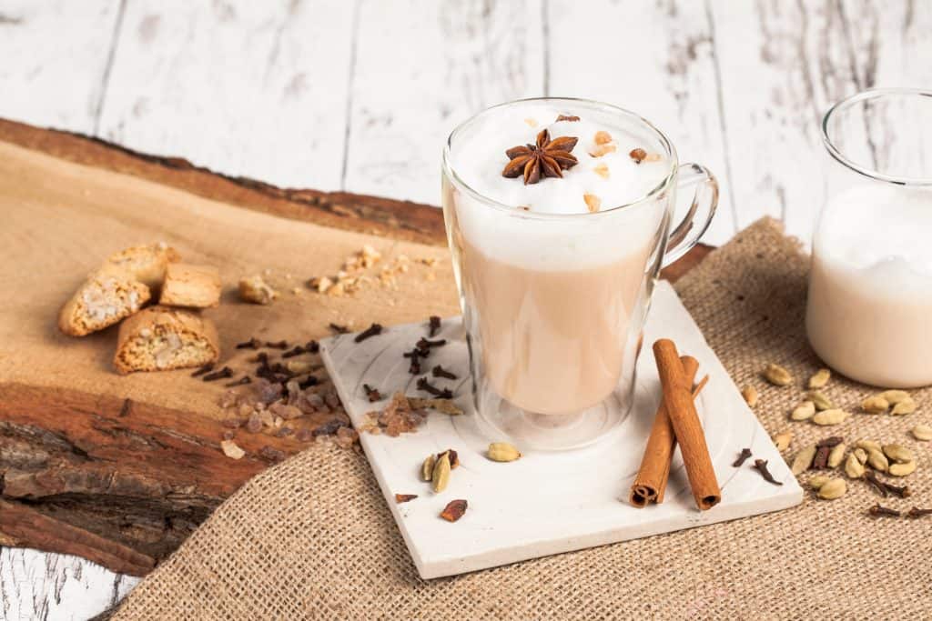 Chai latte in a glass mug sitting on a stone coaster surrounded by spices and cinnamon sticks. 