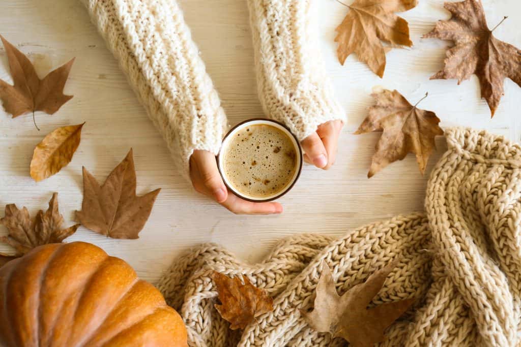 Top view of a woman’s hands holding onto a cup of coffee with leaves and a pumpkin in the background.
