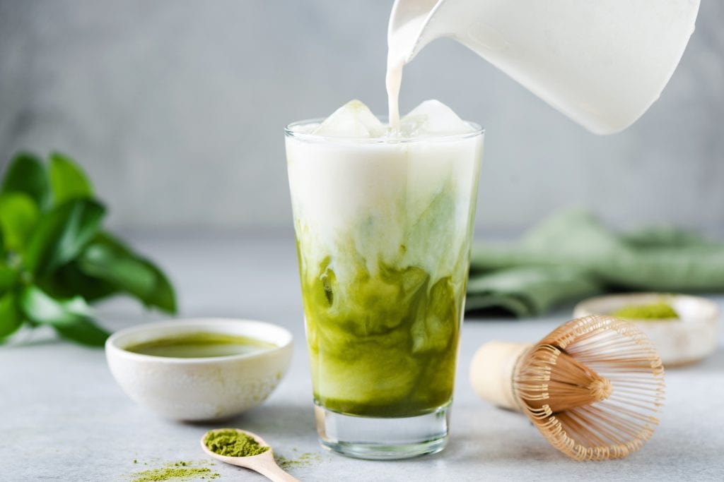 Milk being poured into an iced matcha latte, with a bowl and spoon of matcha nearby.