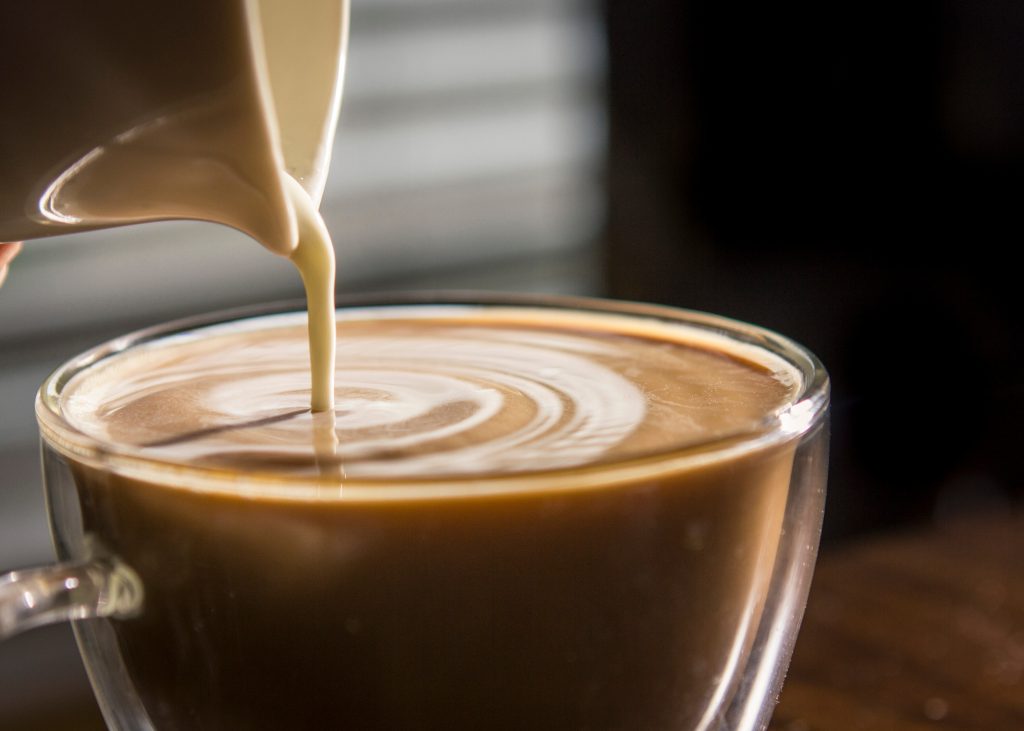 Close up of coffee in a clear glass coffee mug with creamer being poured in.