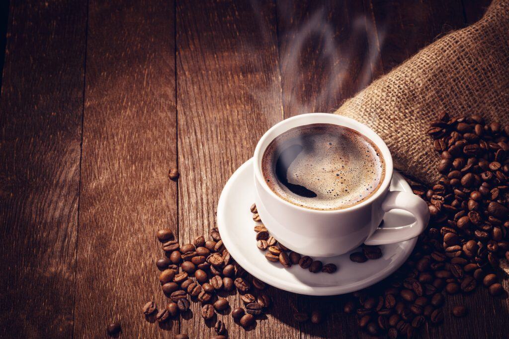 A cup of coffee sitting on a wooden table surrounded by coffee beans.