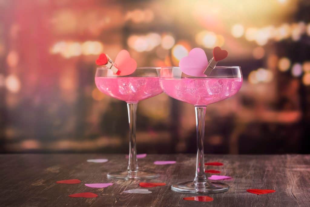 Two pink valentines cocktails with heart garnish on a wooden table with heart confetti scattered on top.