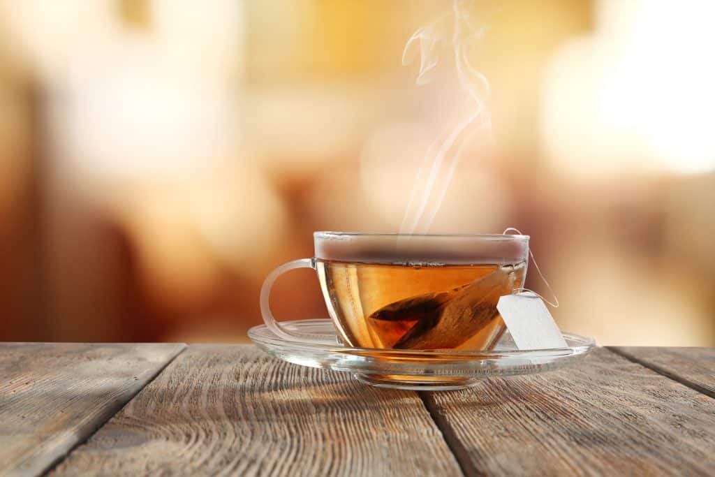Steaming cup of tea in a clear class tea cup and saucer that is sitting on a wooden table.