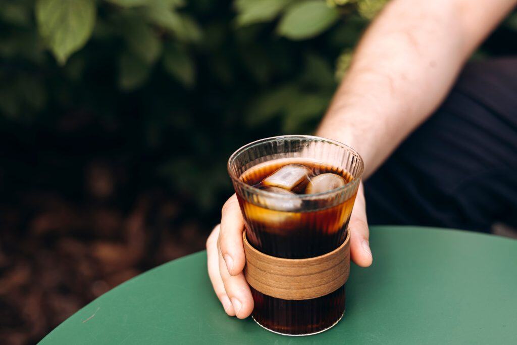 A hand holding onto a glass of cold brew coffee outside on a green table.
